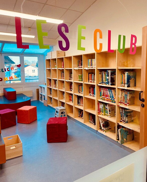 Blick in den Leseclub. Man sieht ein großes Regal mit mehreren Fächern in denen viele Bücher stehen. Es gibt mehrere Sitzwürfel.