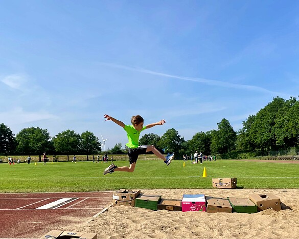 Das Foto zeigt ein Kind in der Luft, dass mit ausgestreckten Armen und Beinen über eine Weitsprunggrube springt